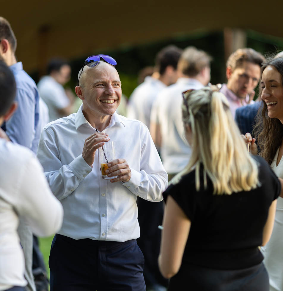 People talking in a circle at an event. There is a man holding a drink and smiling with sunglasses on his head and two women with their back of their heads visible engaged in conversation